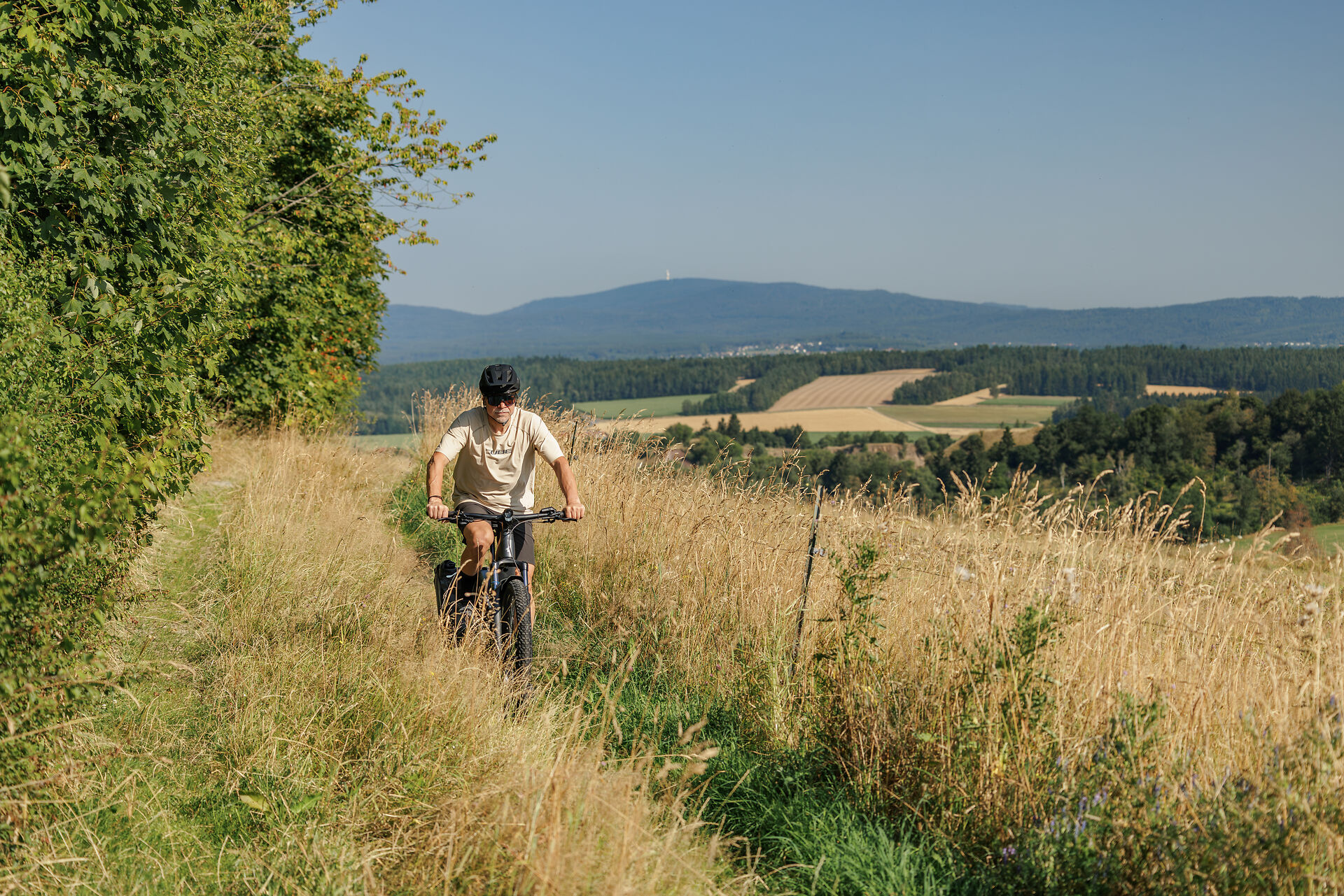 Mountainbiker auf einem schmalen Wiesenpfad über einem weiten Hügelland
